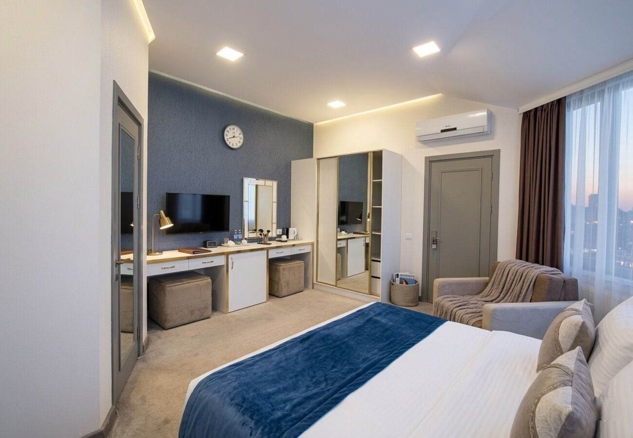 A hotel room with a bed, desk, kitchenette, and a large window with a city view.