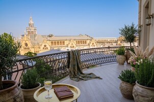 Een balkon met plantenbakken, een tafel met een glas wijn en een boek, en uitzicht op een historisch gebouw met een toren.