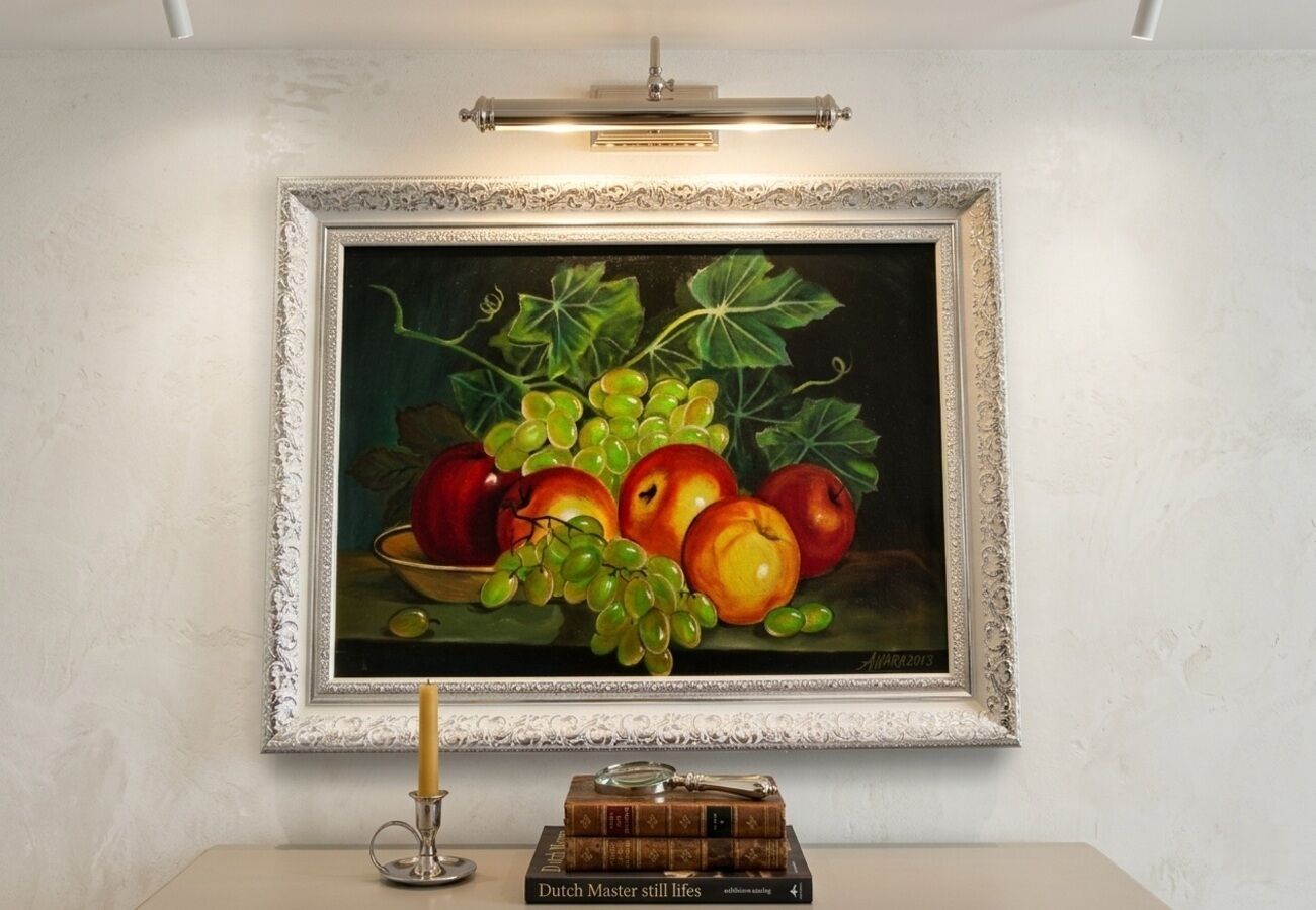 A framed painting of fruits and vegetables on a wall, a candle, and a stack of books.