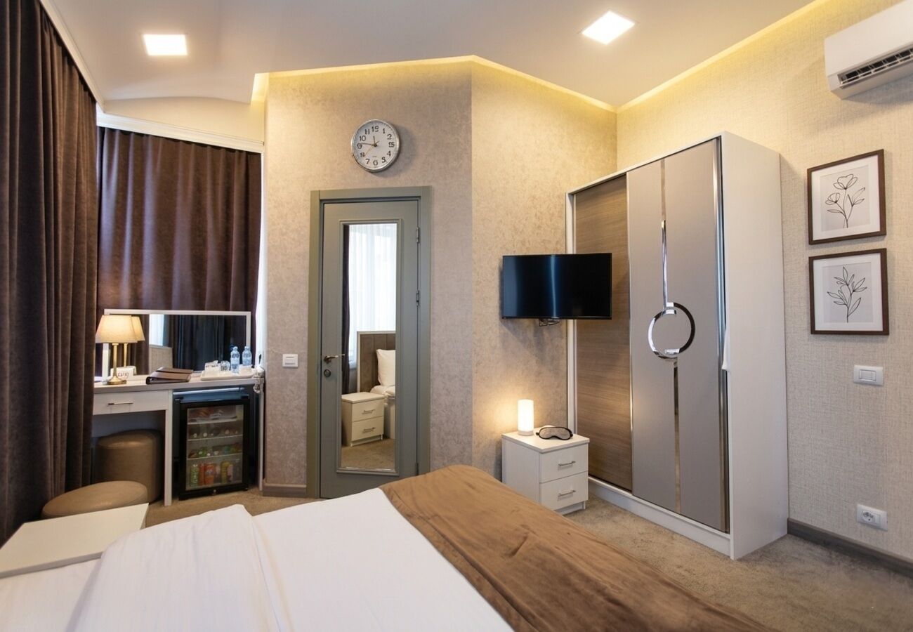 Ett hotellrum med en säng, ett skrivbord med en lampa, en garderob, en tv och en väckarklocka på väggen.