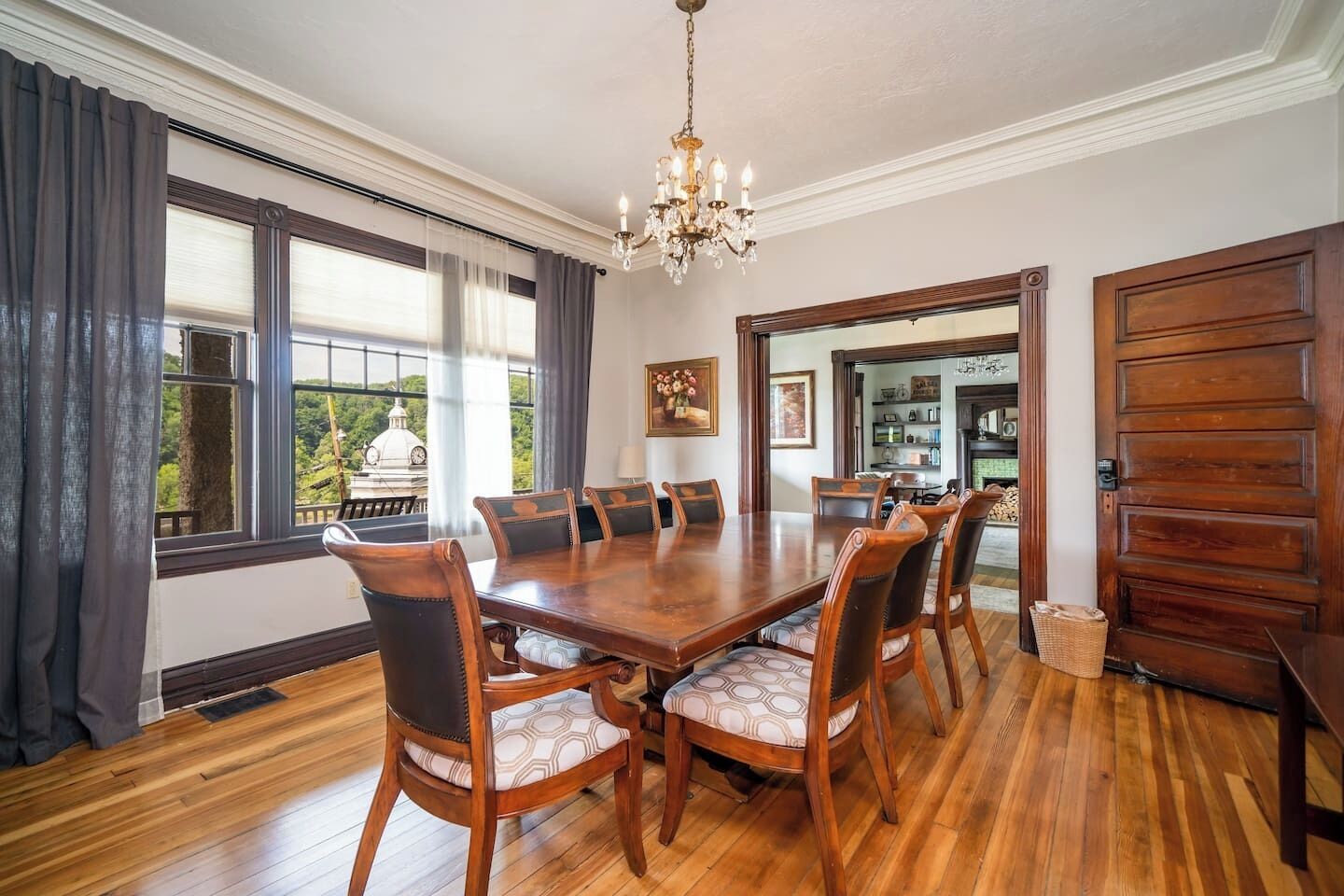 Une salle à manger avec une grande table en bois entourée de chaises, un lustre et de grandes fenêtres ornées de rideaux.