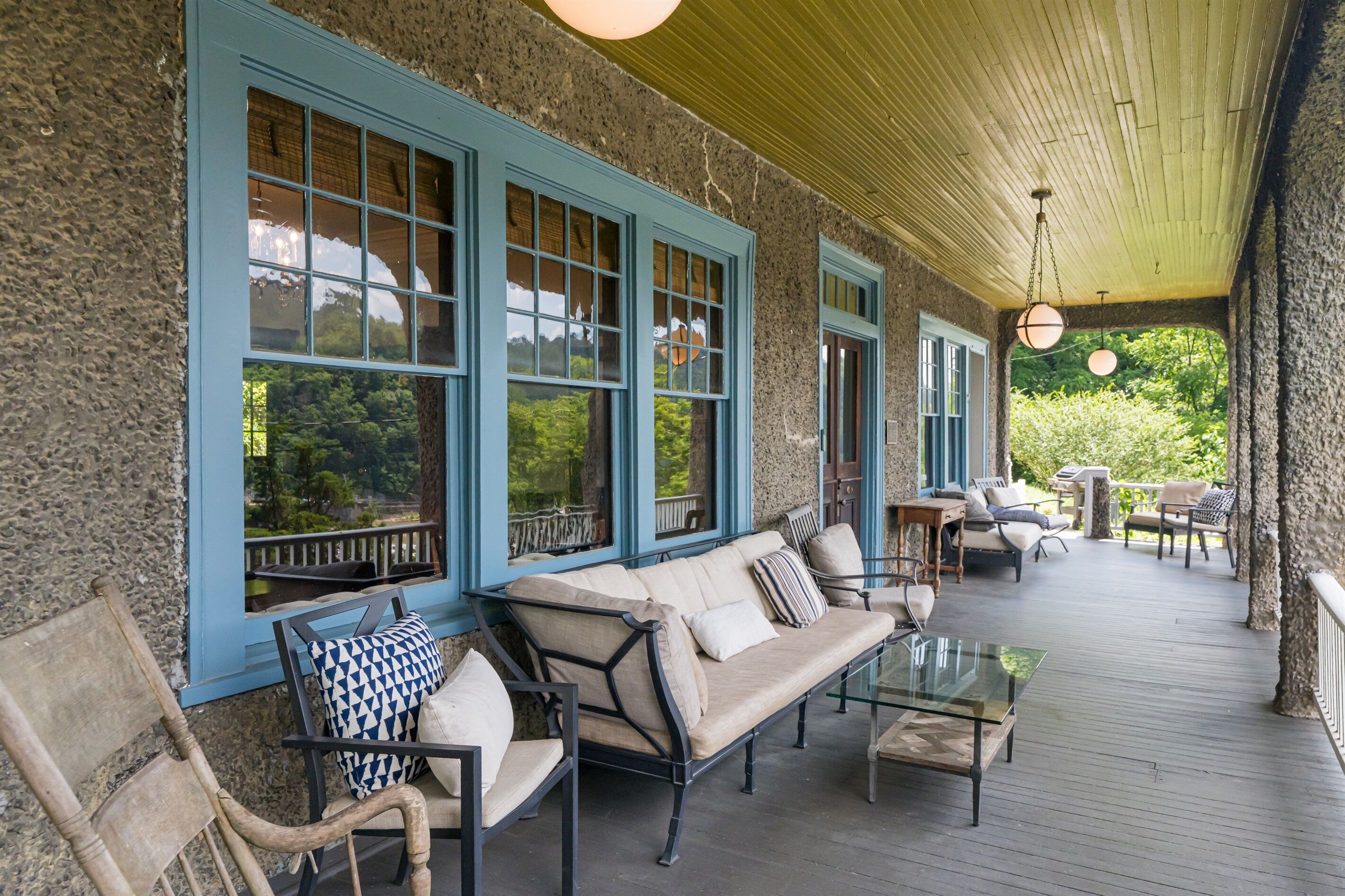 A covered porch with a stone wall, blue-framed windows, a beige sofa, and a glass table.