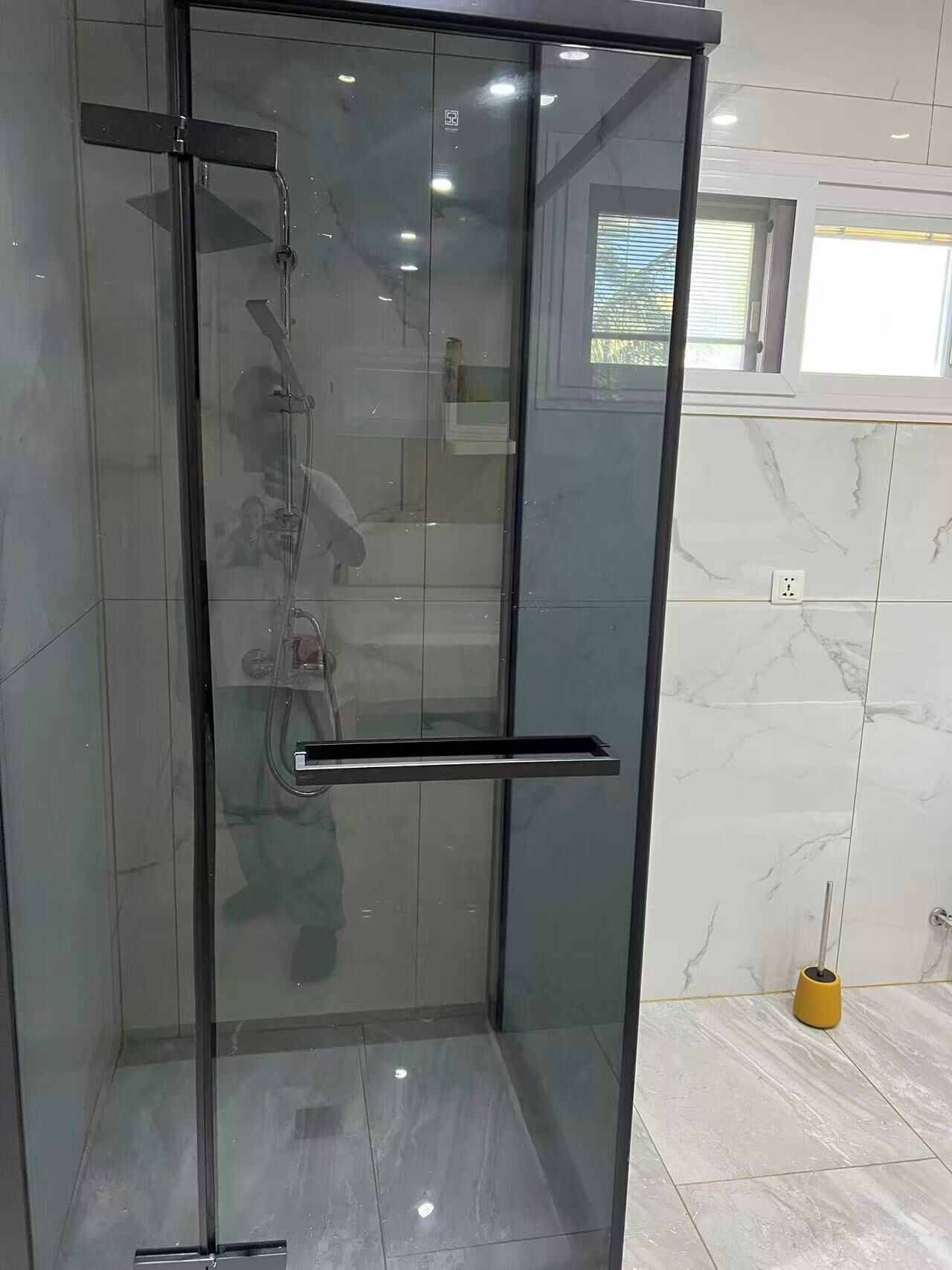 A glass-enclosed shower with a handle, a tiled floor, and a window with blinds.