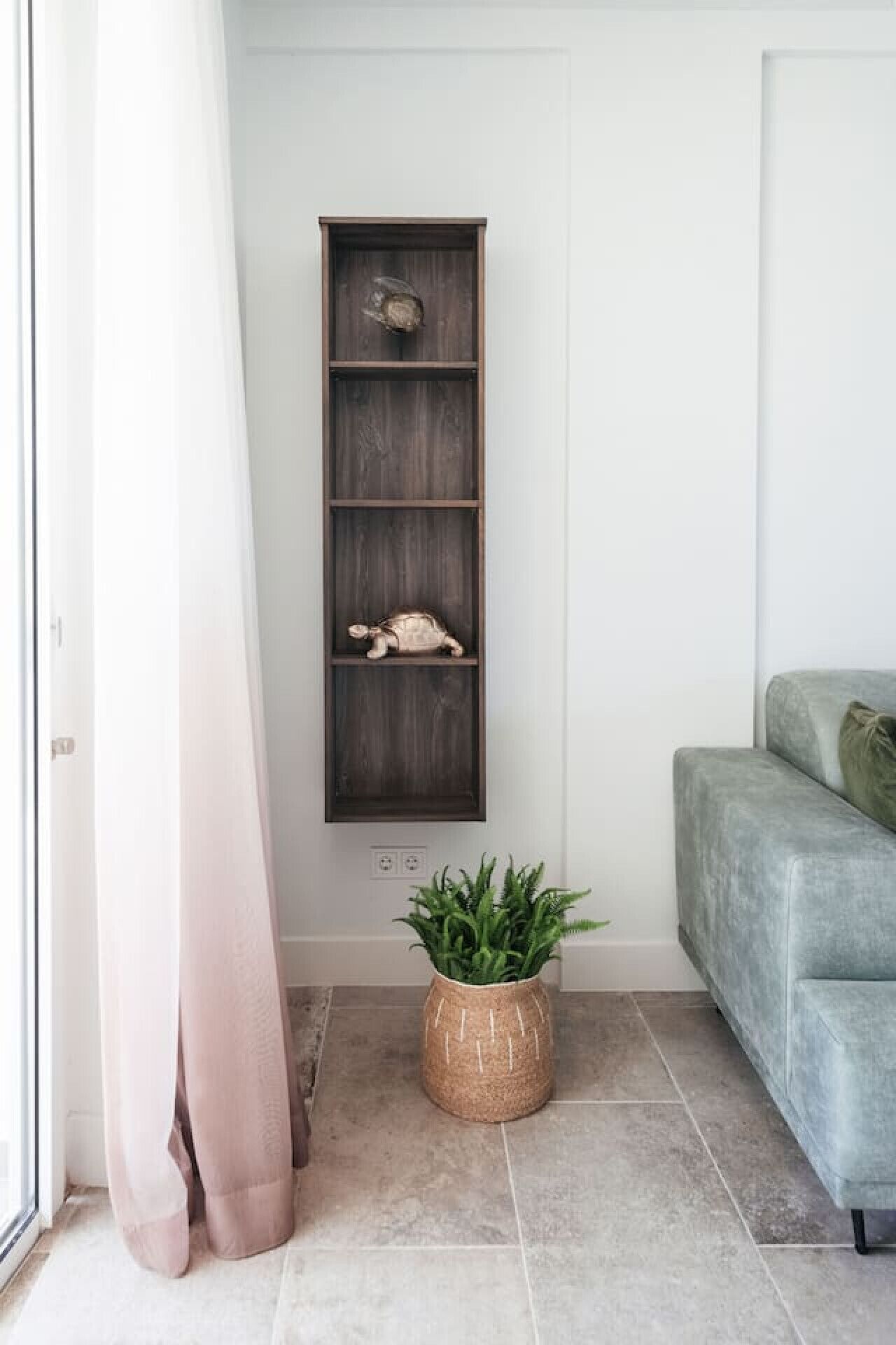 A modern interior with a dark wooden shelf unit against a white wall, a grey sofa, and a woven plant pot on a tiled floor.