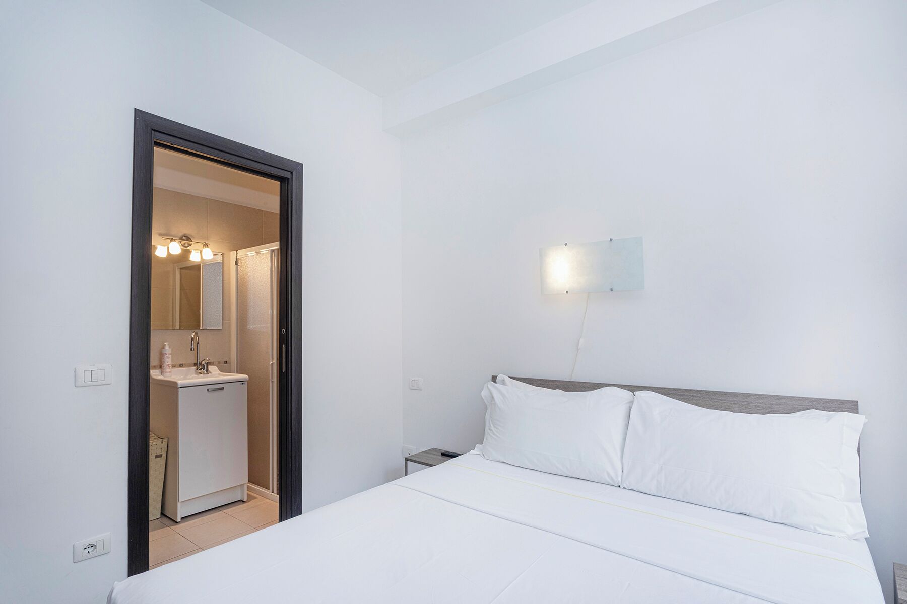 A hotel room with a bed, a nightstand, a wall-mounted light, and a bathroom visible through an open door.