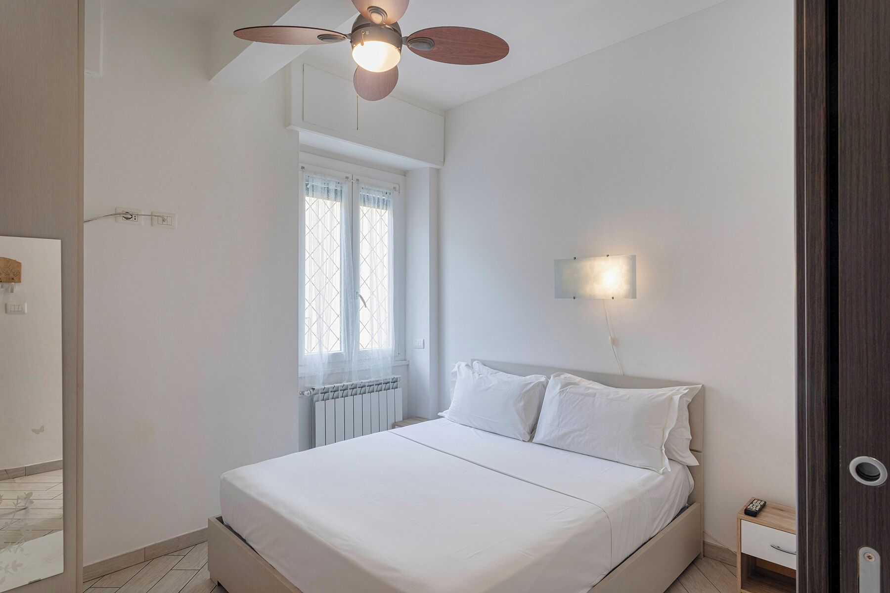 Un dormitorio con una cama, ventilador de techo, ventana con cortinas, lámpara de pared y mesita de noche con control remoto.