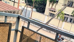 Un balcon doté de balustrades en métal et d’un sol carrelé, donnant sur une rue bordée d’immeubles et d’un palmier.