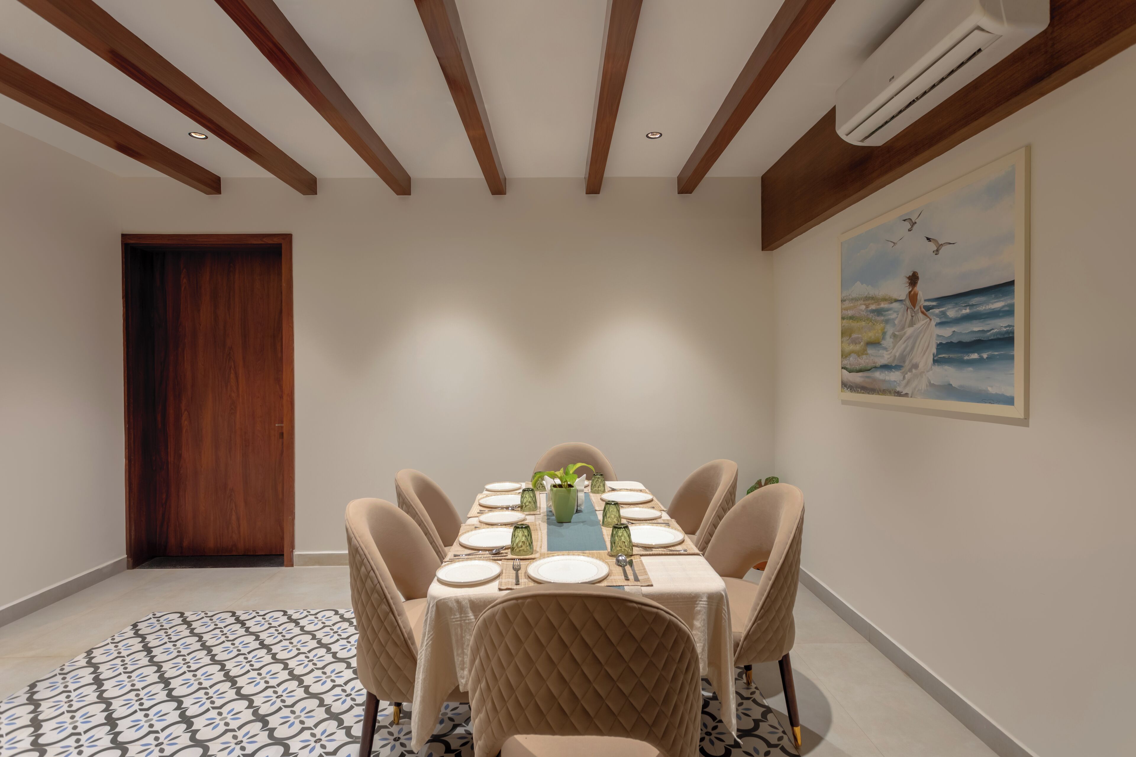 A dining area with a rectangular table set for six, surrounded by upholstered chairs. The room features wooden beams on the ceiling, a large painting on the wall, and a door on the left.