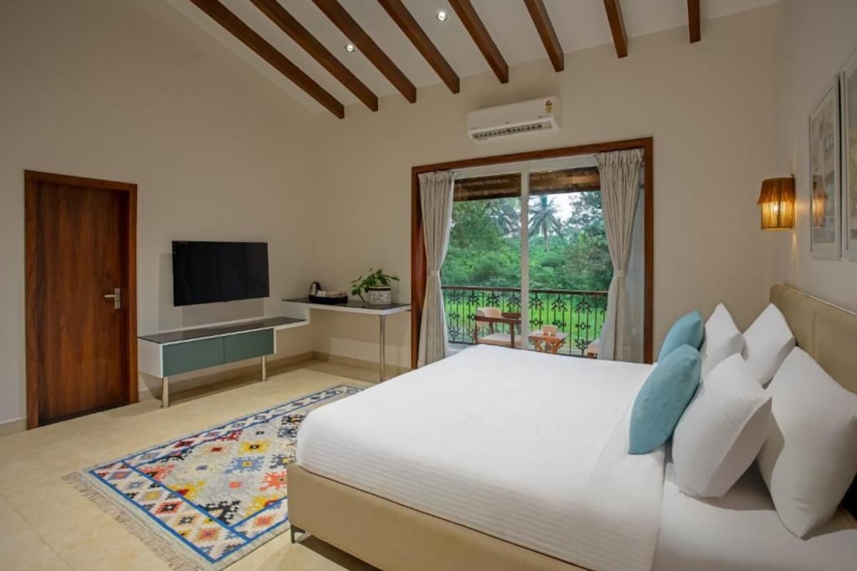 A bedroom with a bed, a TV, a desk, a window with curtains, and a balcony with plants.