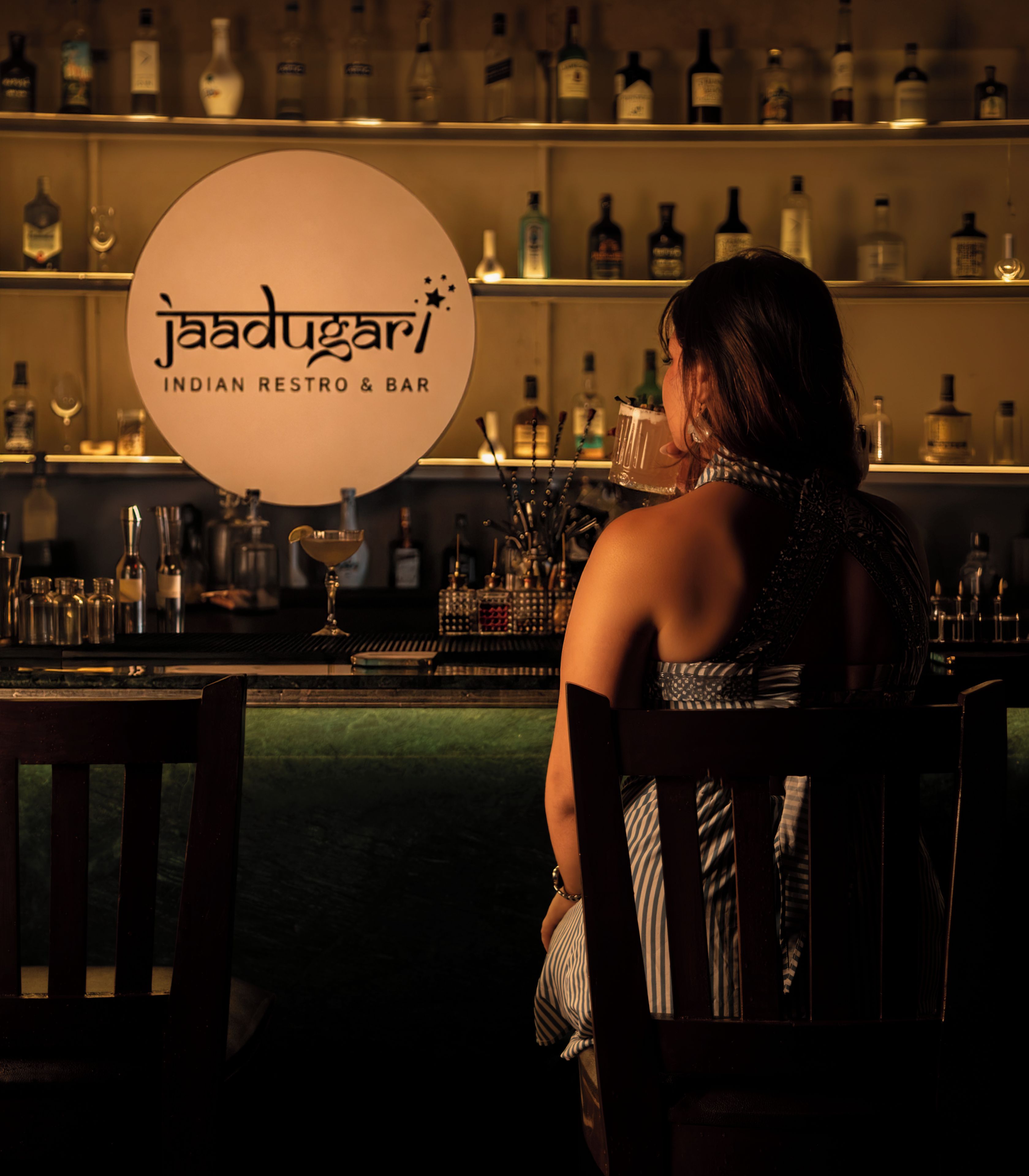 A person seated at a bar with a sign reading 'Jaadughar Indian Restro & Bar' in the background.