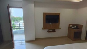 A hotel room with a bed, a TV mounted on the wall, a small wooden desk, and a balcony with a view. - Hotel Raas Inn (Udaipur)
