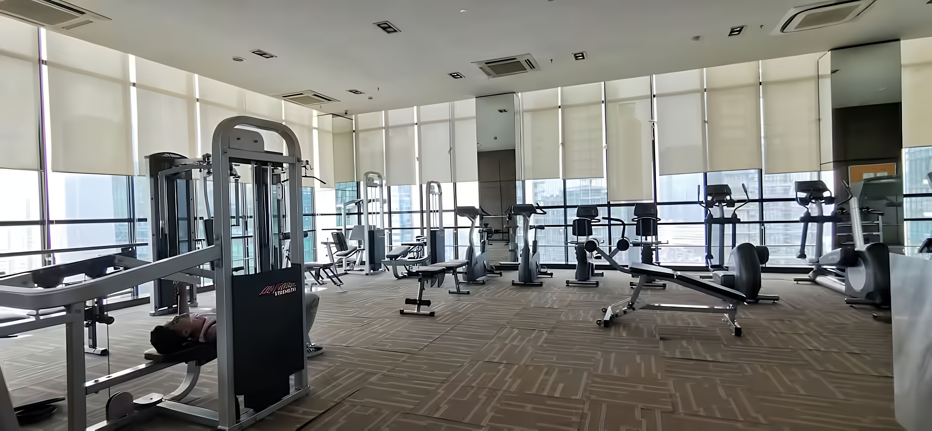 Un gimnasio con diversas máquinas de ejercicio, amplios ventanales y suelo de madera.