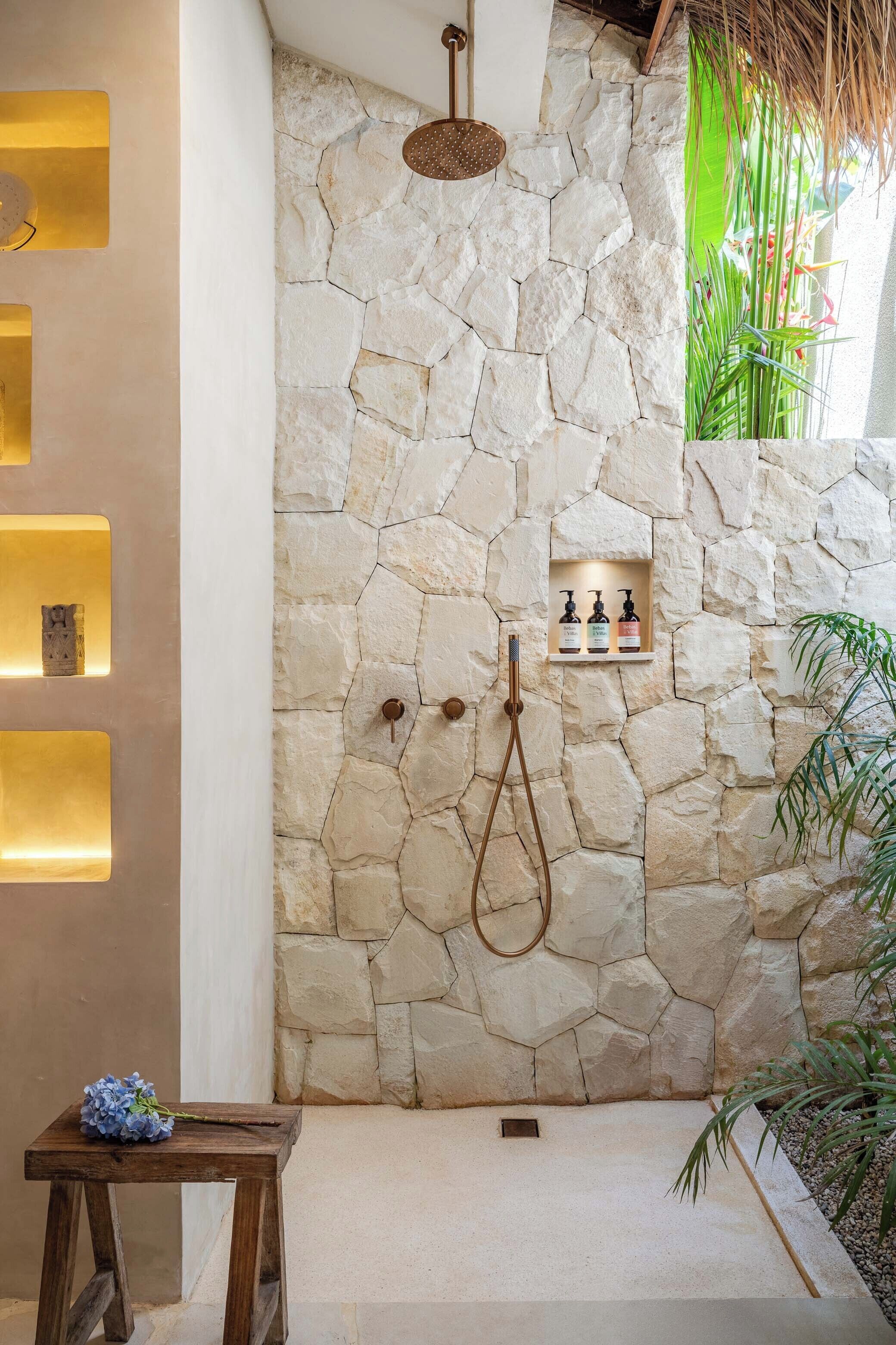 A stone-walled bathroom with a shower, a wooden stool, and a shelf with toiletries.