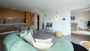 A modern living room with a green sofa, a dining area with chairs, a kitchen with wooden cabinets, and a bed in the background. - Casa Brisa by Acasadascasas (Mafra)