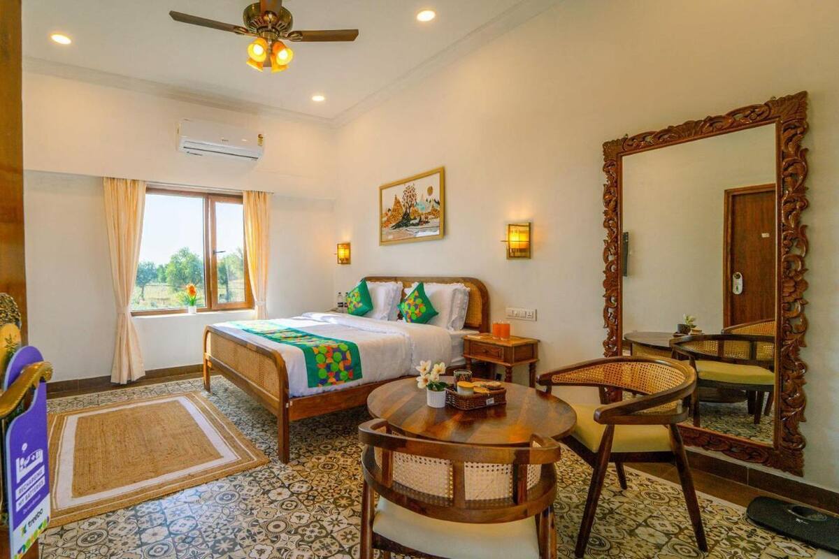 A hotel room with a bed, a round wooden table, a chair, a large mirror, and a window with curtains.