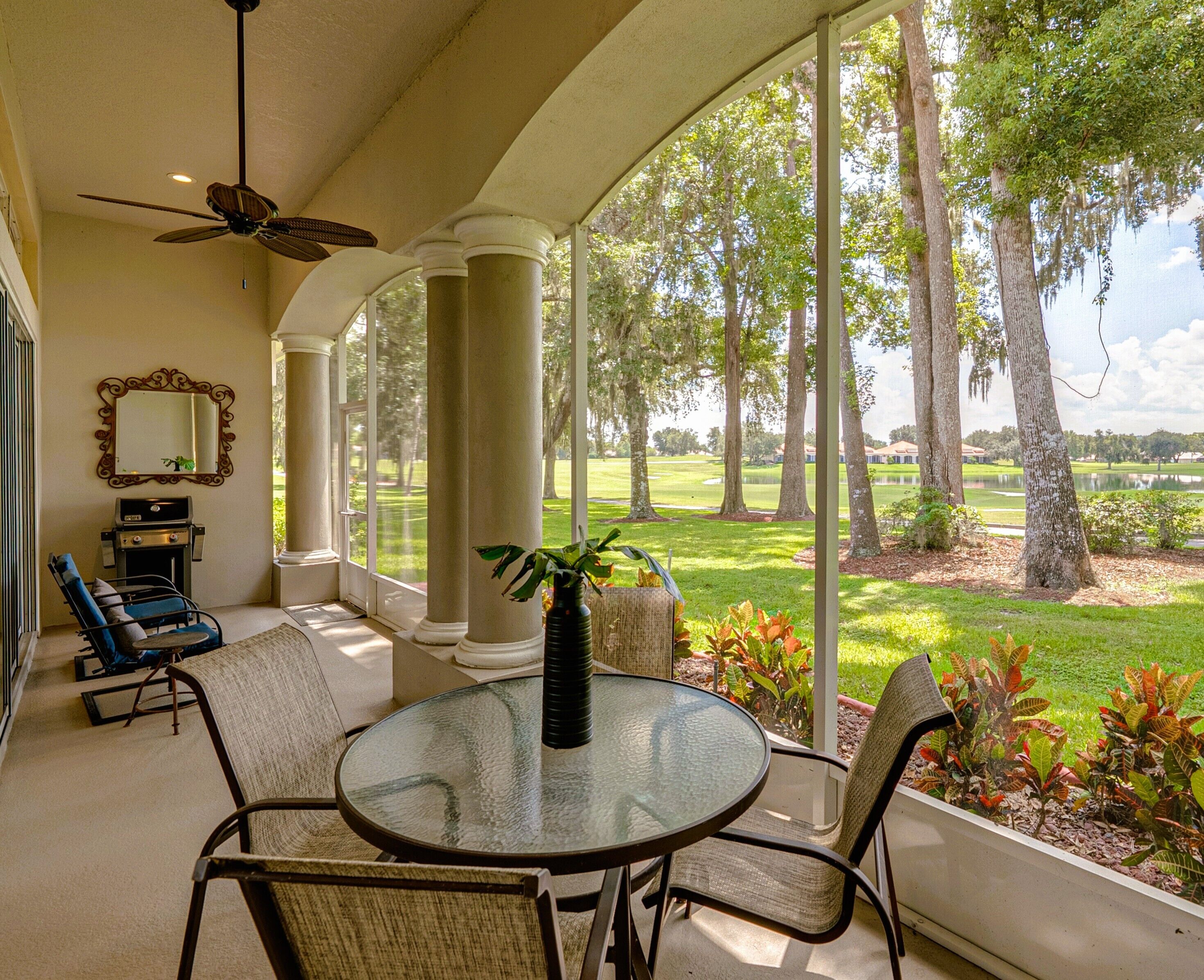 Un patio couvert avec une table et des chaises en verre, un ventilateur de plafond, un barbecue et une vue sur un terrain de golf.