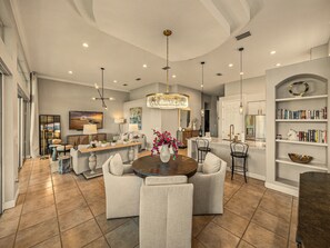 A spacious living room with a round dining table, comfortable seating, a chandelier, and a kitchen area with a bookshelf.