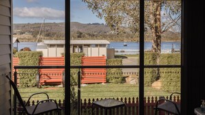 Une vue depuis une fenêtre donnant sur une terrasse en bois, avec deux chaises, et une vue sur un lac où l’on aperçoit des bateaux et un bâtiment en arrière-plan.