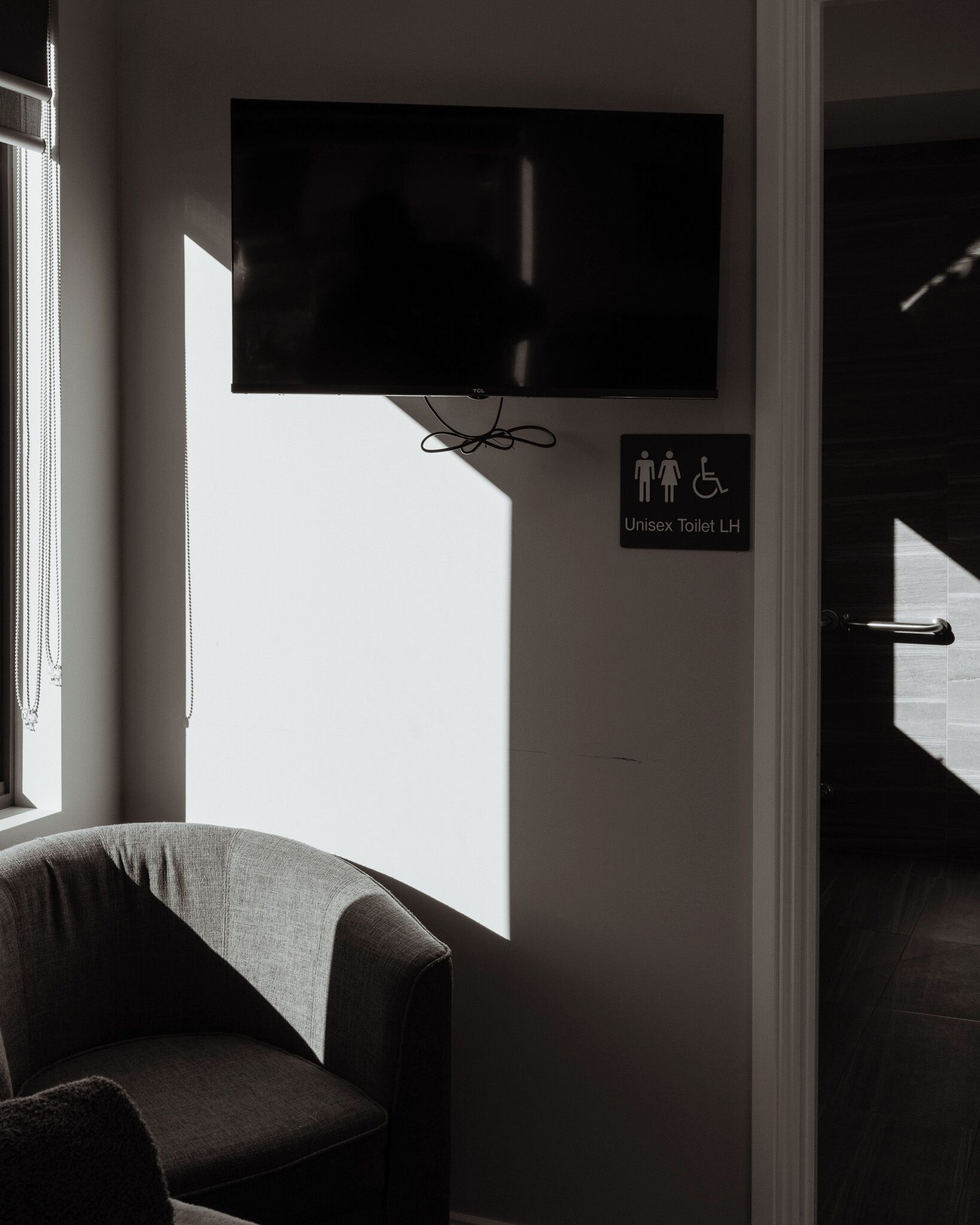 Een kamer met een aan de muur bevestigde televisie, een bordje dat een uniseks toilet aangeeft en een stoel bij een raam.