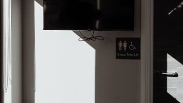 Een kamer met een aan de muur bevestigde televisie, een bordje dat een uniseks toilet aangeeft en een stoel bij een raam.