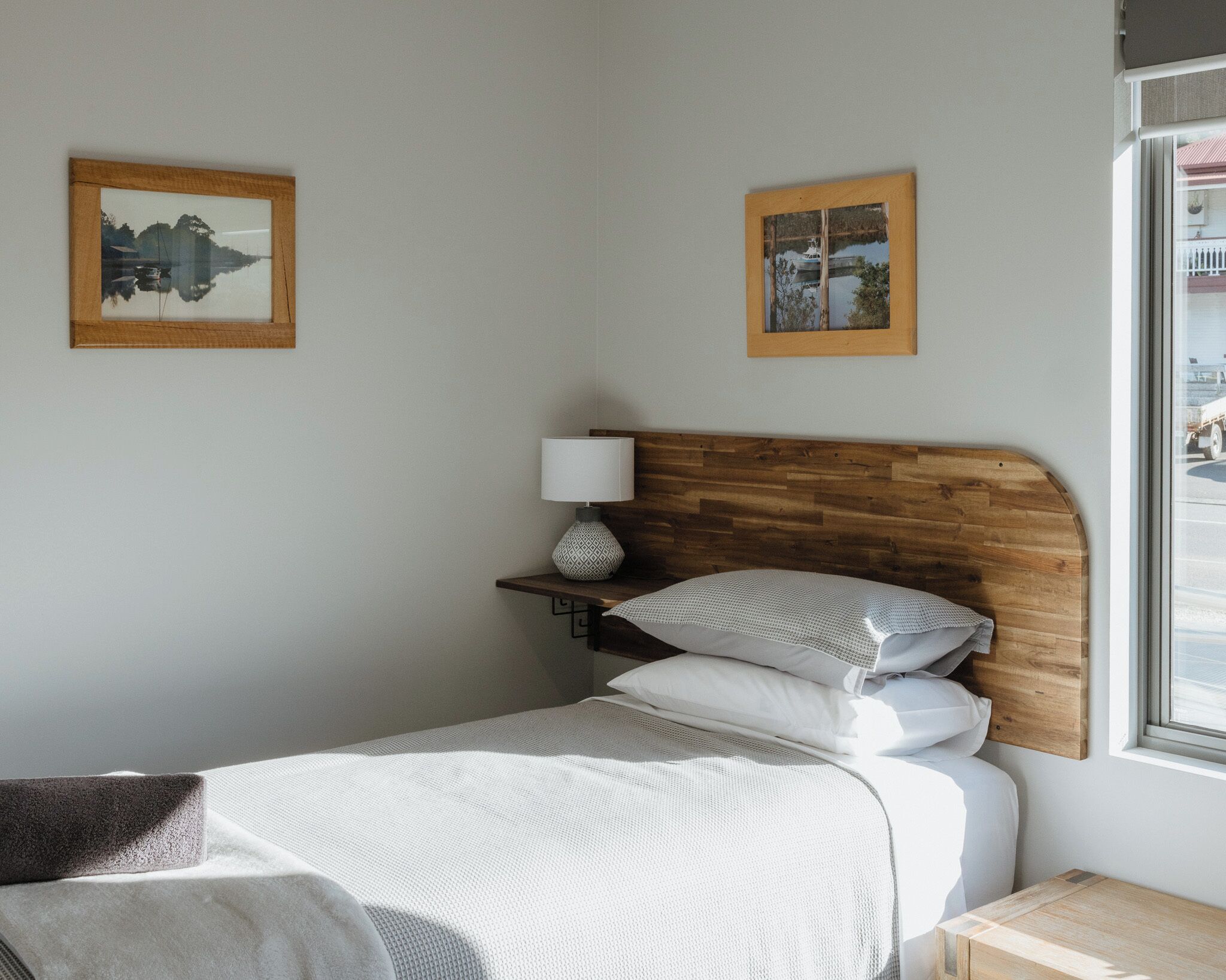 A neatly arranged bedroom with a wooden headboard, a bedside table with a lamp, and two framed pictures on the wall.