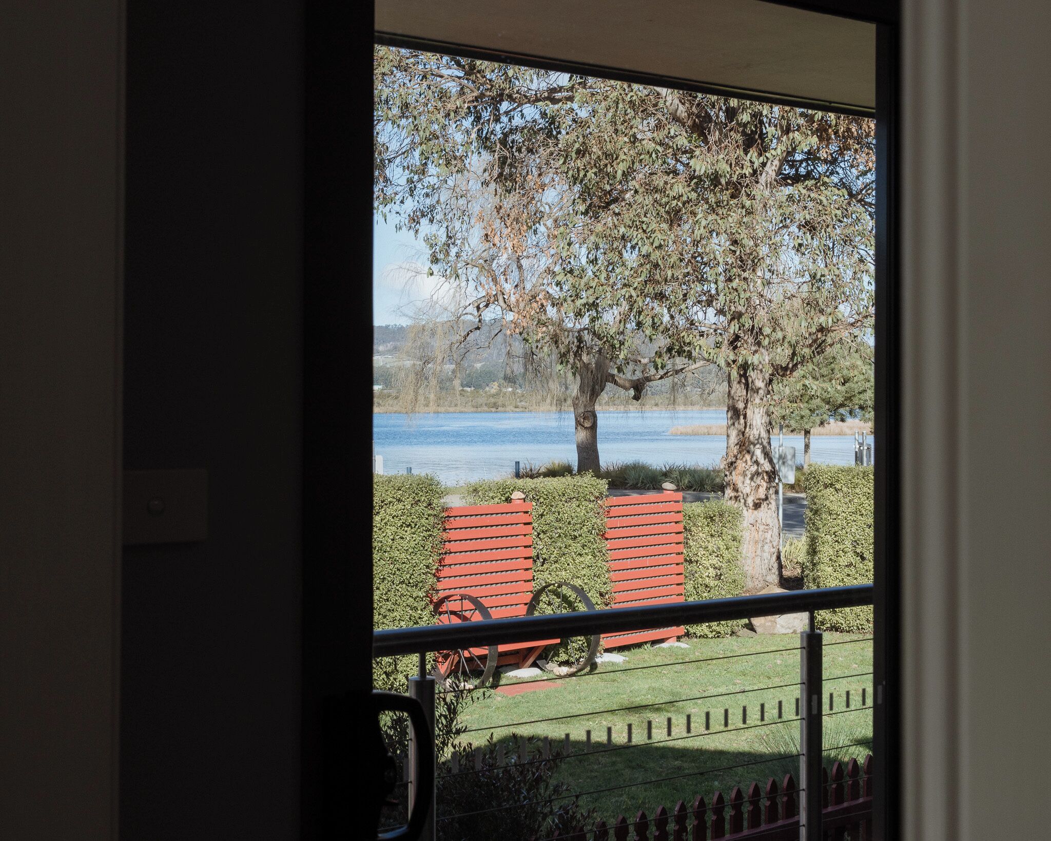 Blick durch ein Fenster in einen Raum mit einer Tür, einer roten Bank und einem Baum in einem Garten.