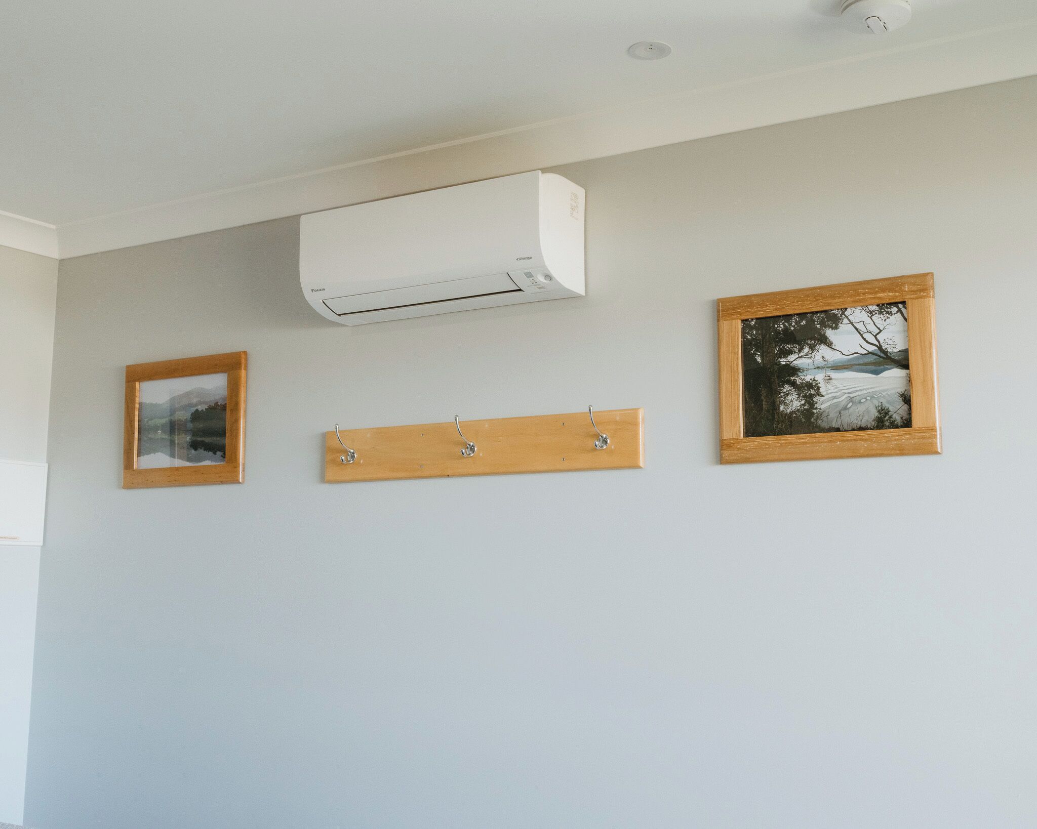 Una habitación con un aire acondicionado blanco instalado en la pared, un estante de madera con ganchos y dos cuadros enmarcados.