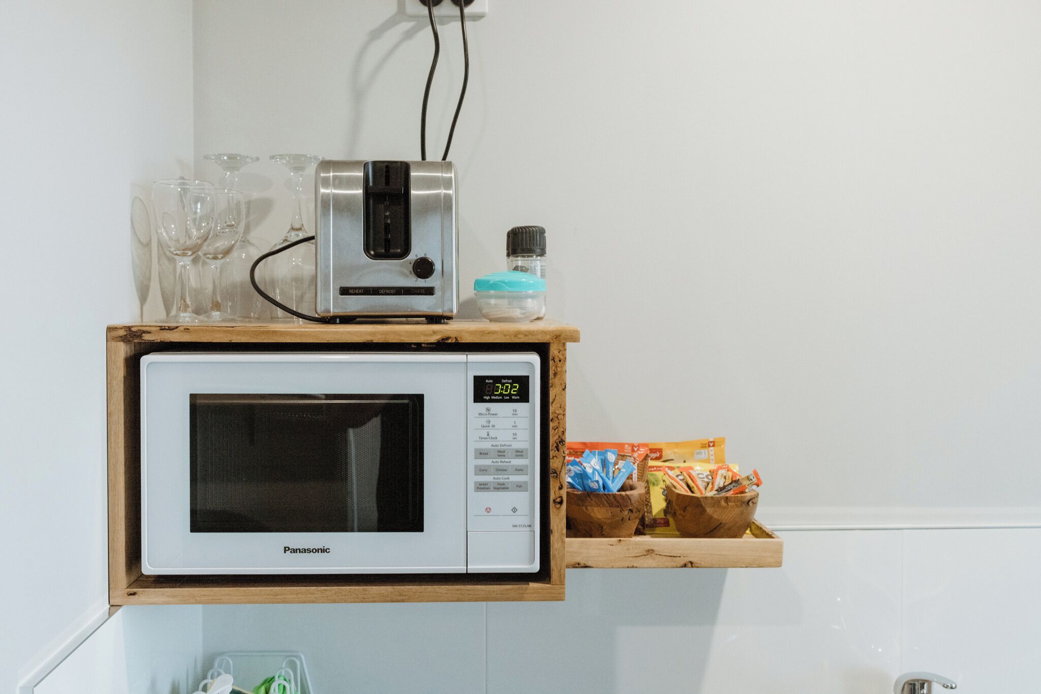 A kitchen with a microwave oven on a wooden shelf, a small appliance on top, and a box of snacks.