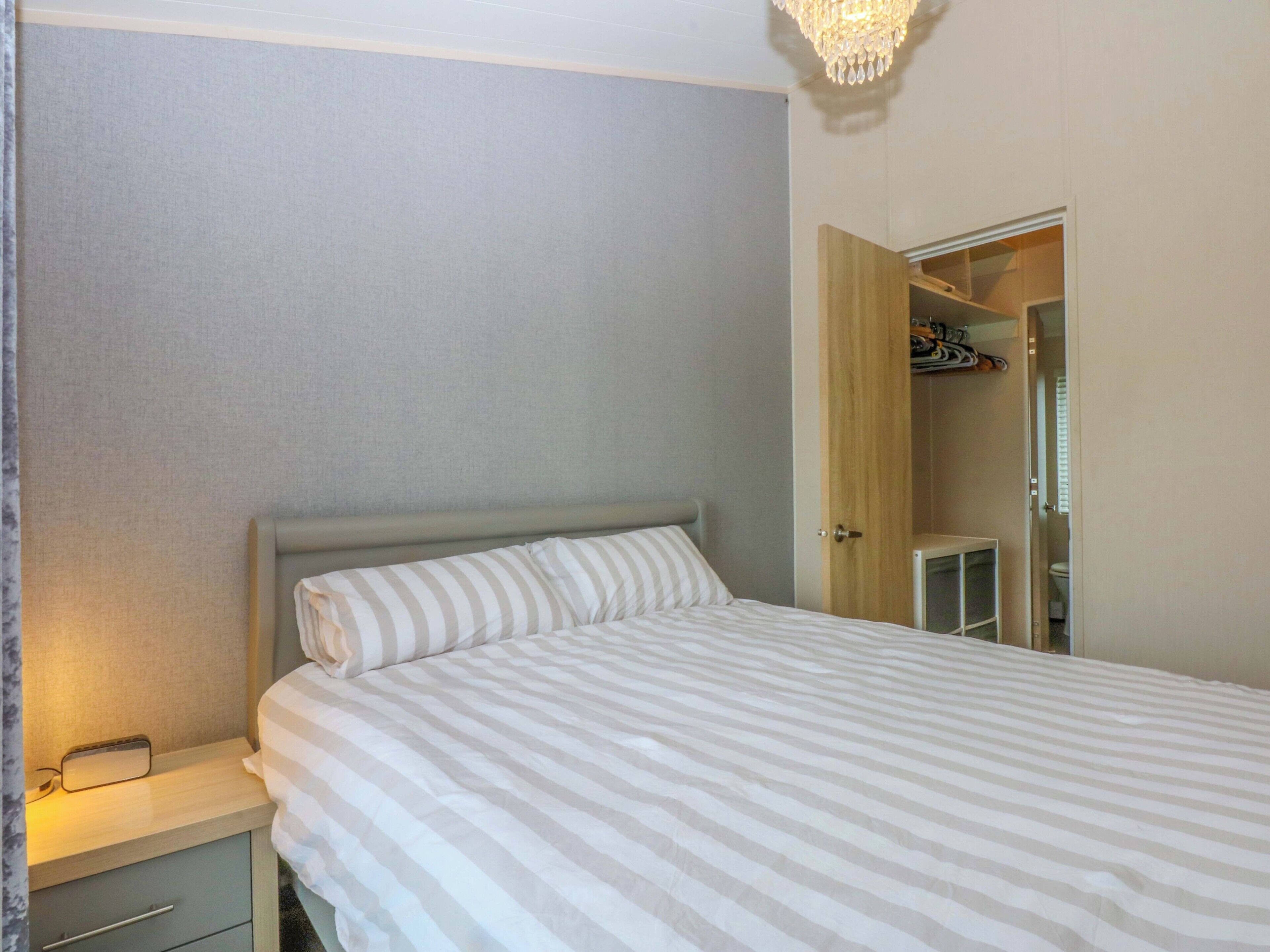 A neatly made bed with striped bedding, a bedside table with a lamp, and a wardrobe with hanging clothes.