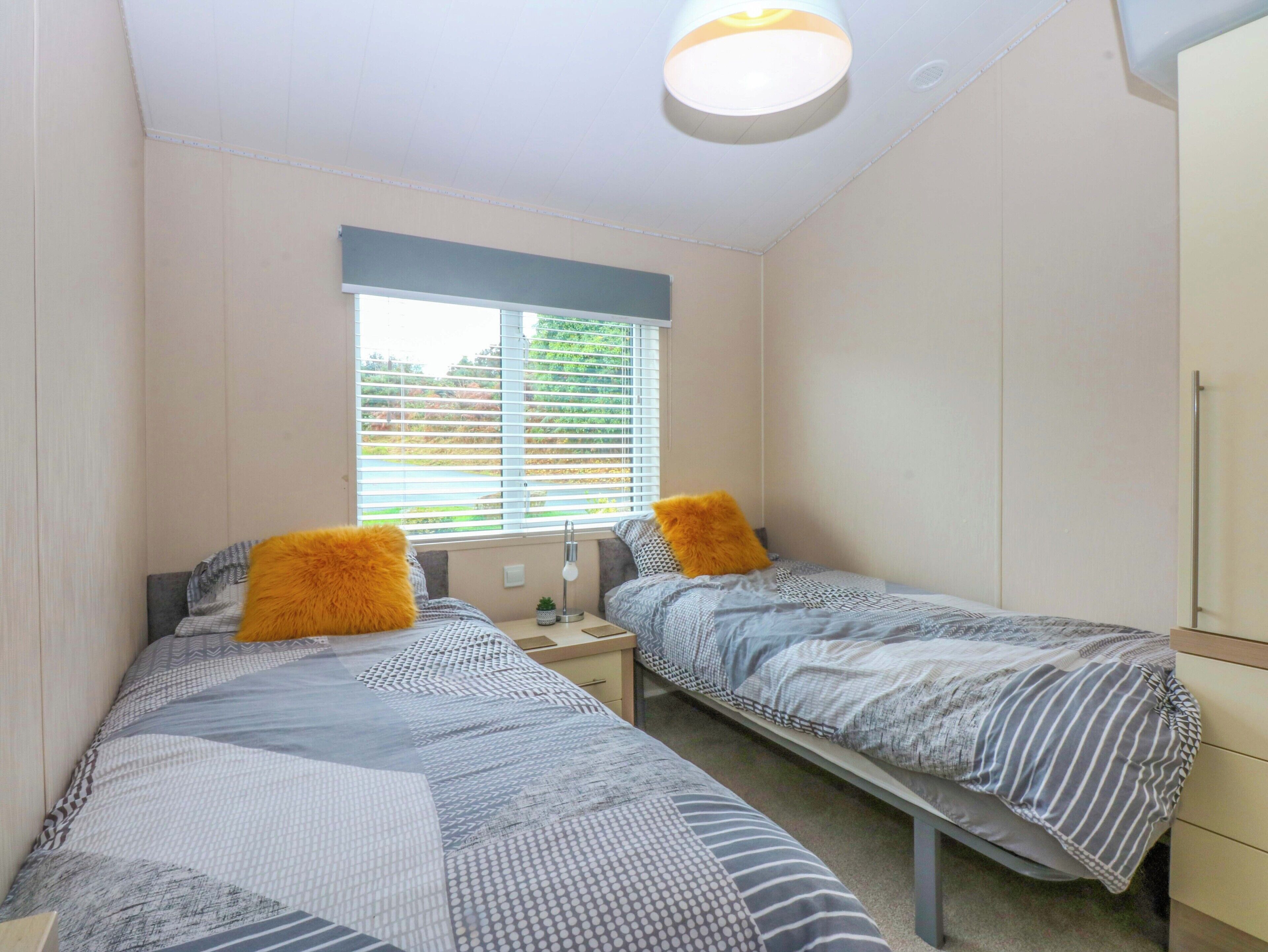 A bedroom with two single beds, a window with blinds, a bedside table with a lamp, and a wardrobe.