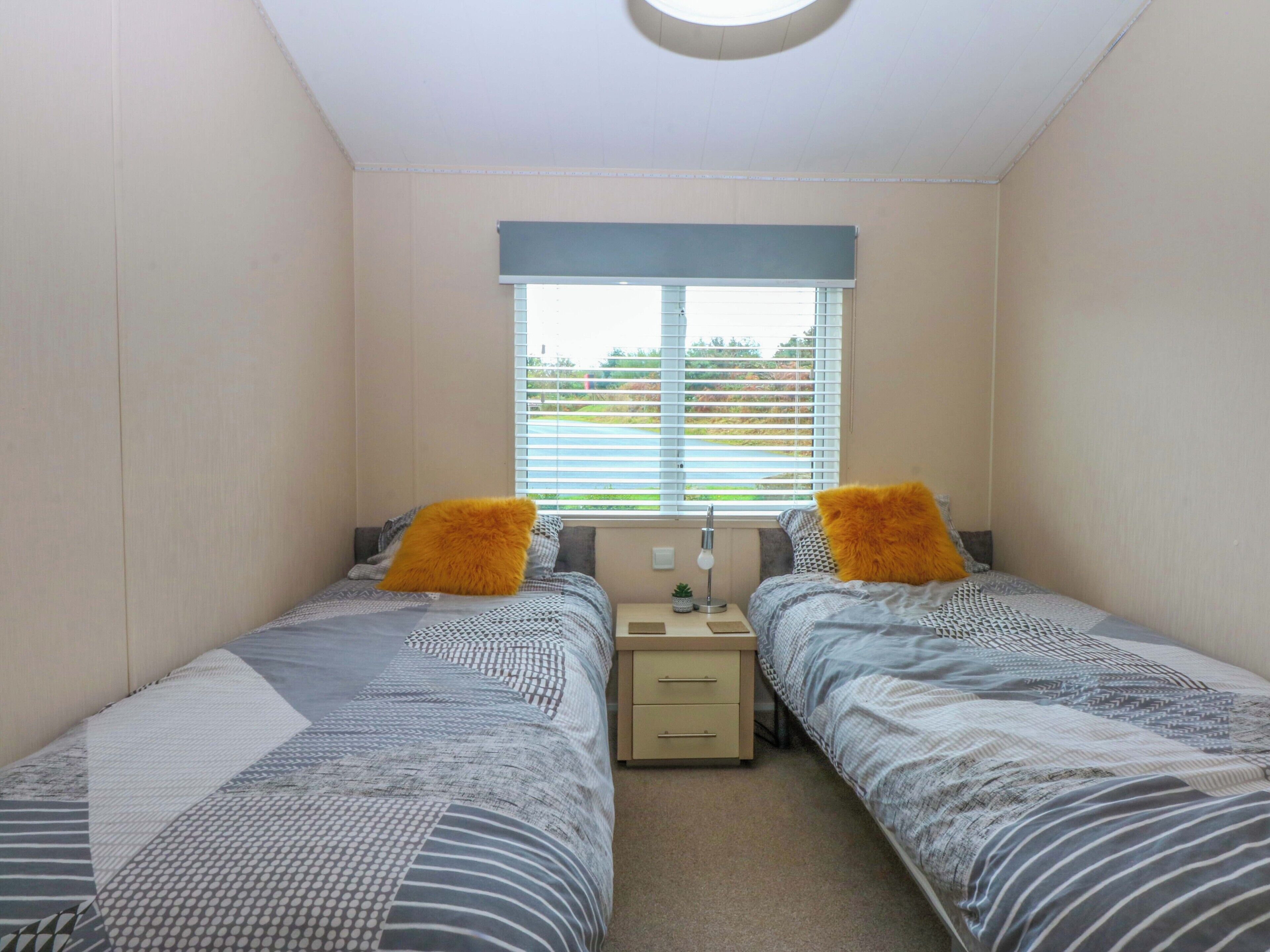 A bedroom with two beds, a window with blinds, a bedside table with a lamp, and a small potted plant.
