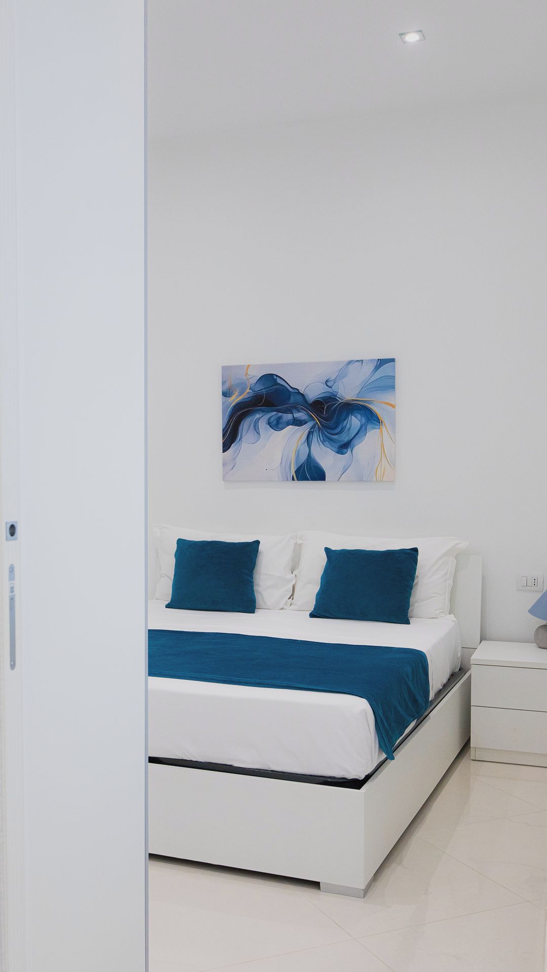 A modern bedroom with a white bed, blue bedding, and a large abstract painting on the wall.