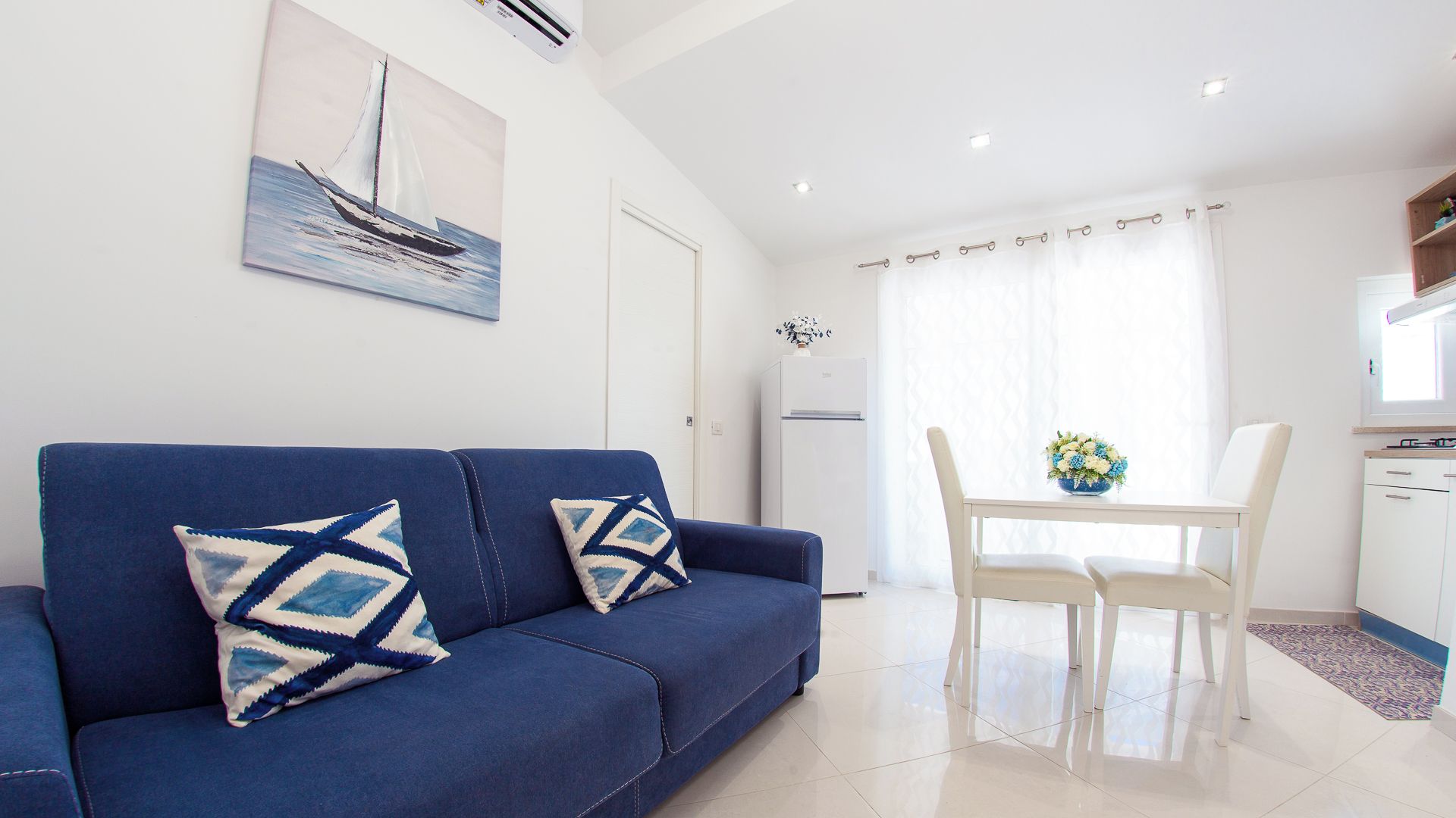 A modern living room with a blue sofa, two patterned pillows, a dining table with chairs, a refrigerator, and a painting of a sailboat on the wall.