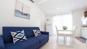A modern living room with a blue sofa, two patterned pillows, a dining table with chairs, a refrigerator, and a painting of a sailboat on the wall. - LA PERLA AZZURRA (Gaeta)