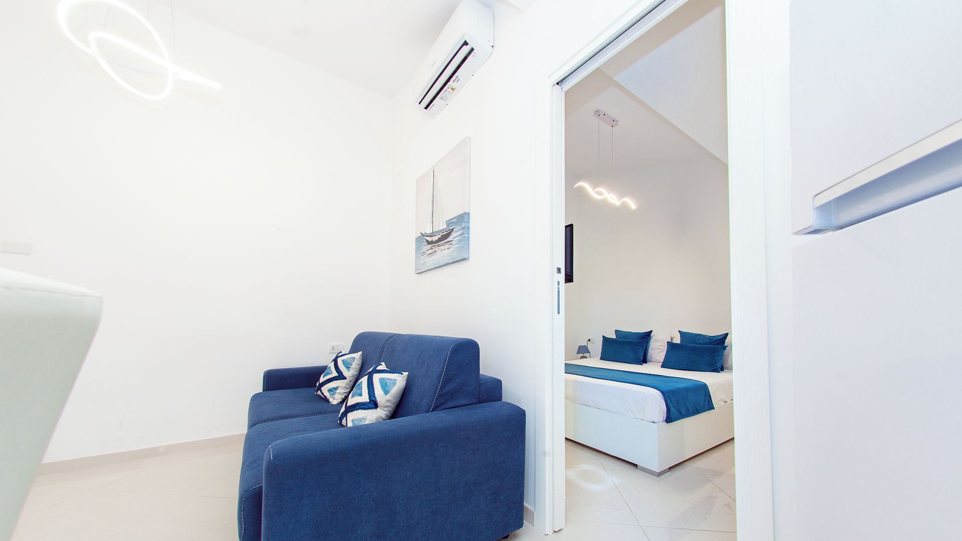 Una habitación moderna con un sofá azul, una cama con ropa de cama azul y una pared blanca con un cuadro enmarcado.