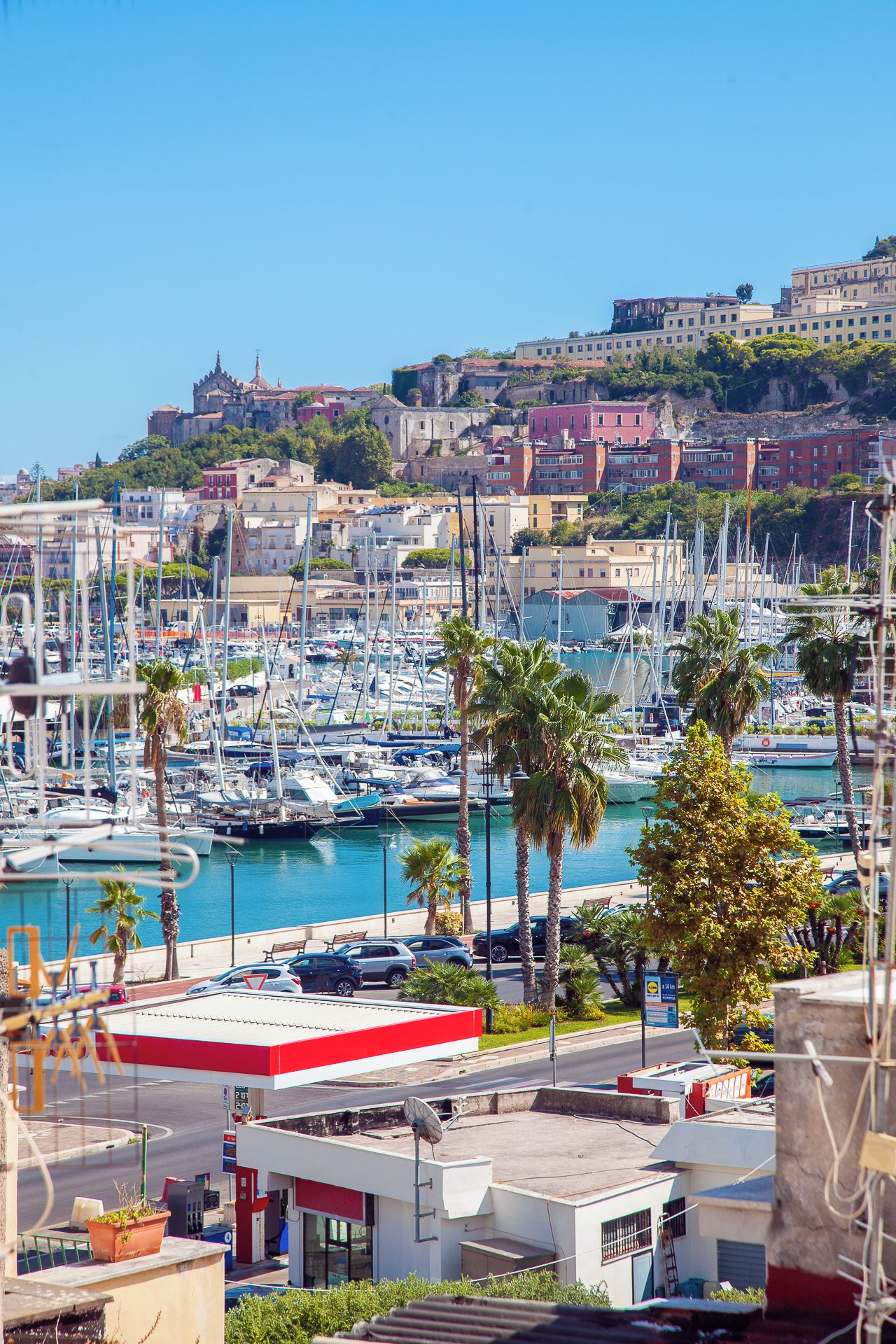 A marina with numerous boats docked, a gas station, and a view of historic buildings in the background.
