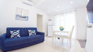 Een moderne woonkamer met een blauwe bank, een eettafel met stoelen, en een keuken met een koelkast.
