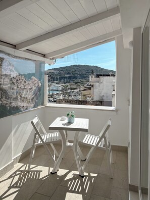 A balcony with a white table and chairs, a view of a marina, and a cityscape in the background. - LA PERLA AZZURRA (Gaeta)
