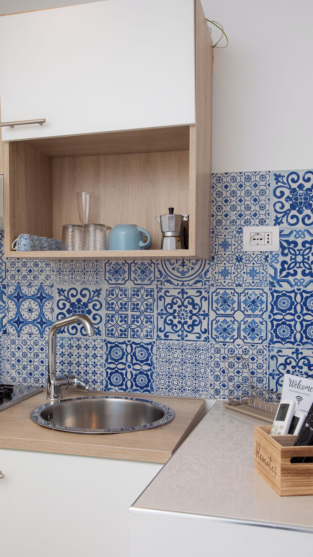 A kitchen with a stainless steel sink, a wooden cabinet above it, and blue patterned tiles on the wall.