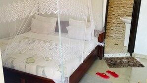 A neatly made bed with a canopy, a small bedside table, a rug, and a pair of slippers on the floor. - Fanad Hotel (Gisenyi)