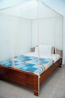Una cama individual con estructura de madera y colcha azul estampada, un almohadón blanco y un dosel con diseño de encaje.