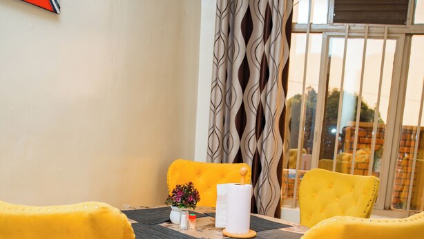 A dining area with a table set for four, yellow upholstered chairs, a floral tablecloth, and a view through a window with curtains. - Fanad Hotel (Gisenyi)
