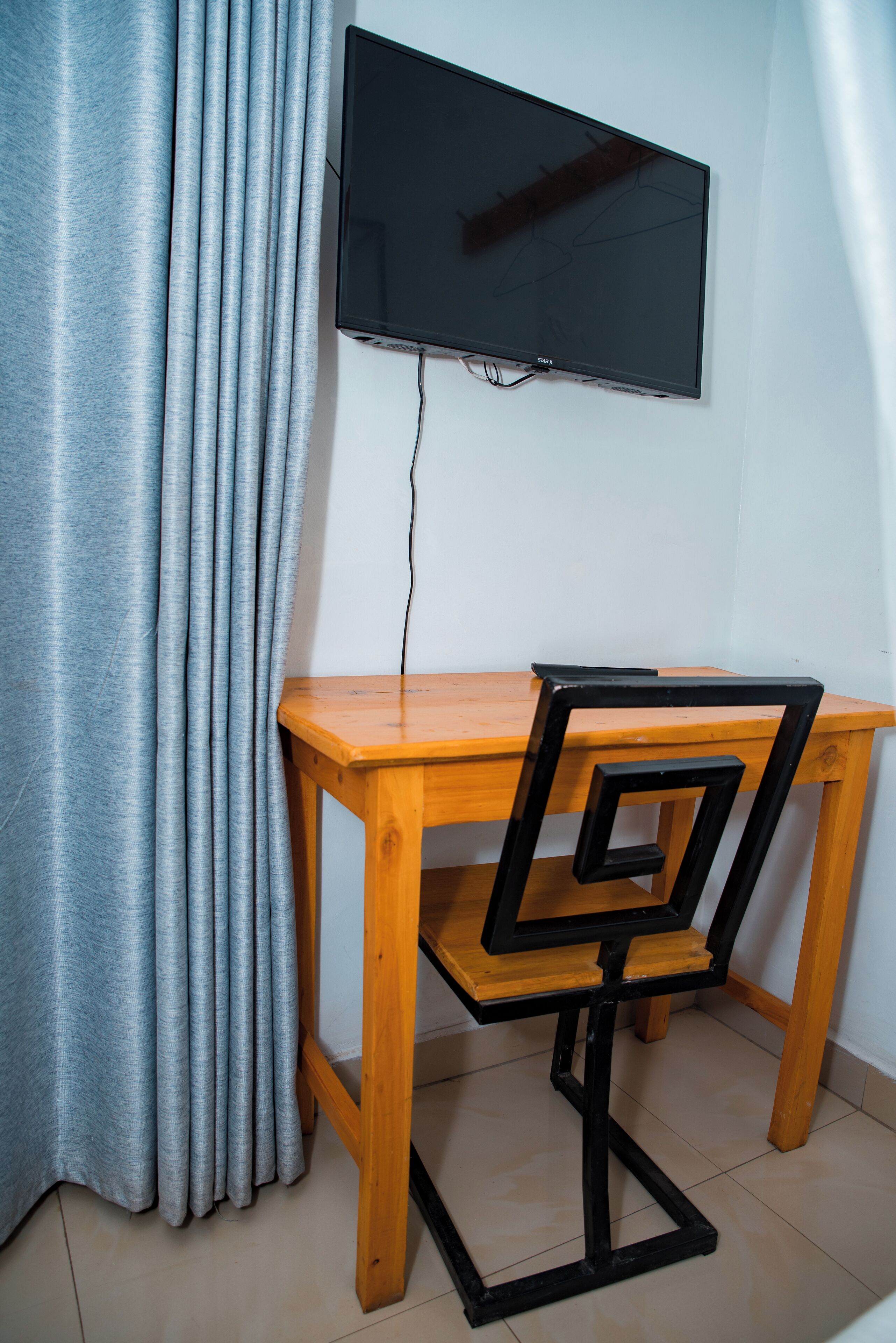 Een houten bureau met een zwarte stoel, een aan de muur bevestigde televisie en een grijze gordijn.