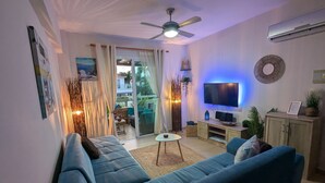 A living room with a blue sofa, a TV, and a ceiling fan. - Andros Apt 2 Bed Free Wi-fi Safe Complex Pool (Paralimni)