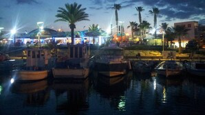 A marina at dusk with boats docked, palm trees, and buildings in the background. - Andros Apt 2 Bed Free Wi-fi Safe Complex Pool (Paralimni)