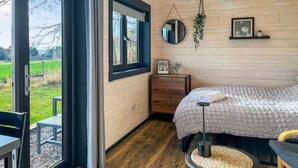 A bedroom with a large window, a bed, a bedside table, a mirror, and a hanging plant. - Barn Owl Cabin at Redroofs - Pet Friendly! (Forfar)