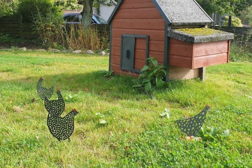 A wooden chicken coop with a sloped roof and a small door, surrounded by grass and plants.
