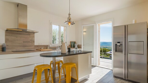 A modern kitchen with a central island, yellow stools, stainless steel appliances, and a view through an open door. - Luxury Villa – Panoramic Views of Mountains, Wi-Fi, and Air Conditioning (Volx)