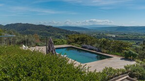 A rectangular pool with a wooden deck, surrounded by lush greenery and a view of distant hills. - Luxury Villa – Panoramic Views of Mountains, Wi-Fi, and Air Conditioning (Volx)