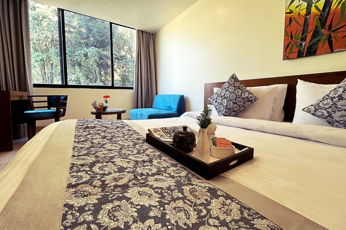 A hotel room with a large bed, a blue armchair, a small table with a tray, and a window with a forest view.